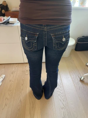 Mörkblå bootcut jeans från true religion - Säljer ett par trendiga mörkblå bootcut jeans från true religion med kontrastsömmar och stora fickor bak med dekorativa knappar och broderade fickoroch är tillverkade i denim med lätt stretch för skön passform. Skäljer då jag inte gillar hur de sitter på mig vilket är synd då jag älskar hur de ser ut. Jag är 166 cm lång.(byxorna är uppsydda 