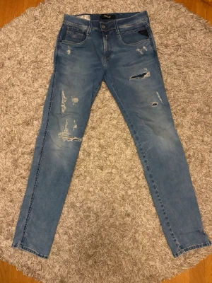 Ljusblå slitna skinny jeans - Säljer ett par ljusblå Replay ANBASS hyperflex jeans med slitningar och hål på framsidan. Jeansen har fem fickor, normal midja och klassisk dragkedja med knapp. Materialet är denim med stretch för extra komfort. Perfekta för en avslappnad och trendig look