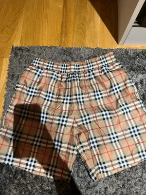Beige rutiga shorts med resårmidja - Snygga beige shorts med klassiskt rutigt mönster i svart, vitt och rött. Shortsen har elastisk midja och sidofickor, perfekta för en avslappnad och trendig look. Tillverkade i ett lätt och bekvämt material som passar bra till sommaren.
