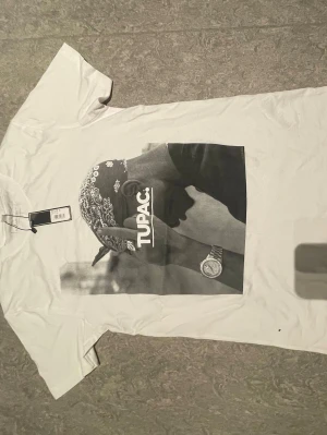 Vit topp med Tupac-tryck - Säljer en vit T-shirt med stort svartvitt tryck av Tupac på framsidan. T-shirten har klassisk passform och korta ärmar. Perfekt för dig som gillar streetwear och vill ha en ikonisk look.