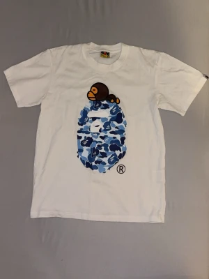 Vit Bape (A bathing ape) T-shirt - Säljer en vit Bape  t-shirt med ett stort blått camomönster i form av ett ansikte på bröstet och en liten apa ovanpå. T-shirten har rund hals och korta ärmar. Perfekt för dig som gillar streetwear och coola prints.