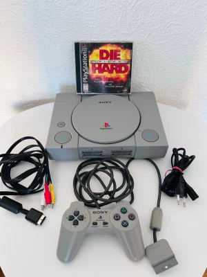 Sony PlayStation 1 med tillbehör - Klassisk grå PlayStation 1 med originalkontroll, minneskort, AV-kablar och strömkabel. Konsolen har rundad form och tydliga PlayStation-loggor. Ett spel, Die Hard, medföljer. Perfekt för dig som gillar retrospel och vill återuppleva gamla favoriter.