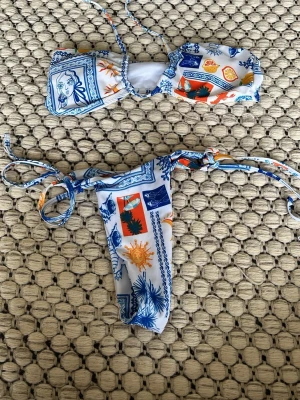 Färgglad bikini med knyt och mönster - Säljer en snygg bikini med bandeau-topp och knyt i sidorna. Bikinin har ett färgstarkt mönster i blått, orange och gult på vit botten, med motiv som sol, ansikten och växter. Perfekt för stranden eller poolen i sommar! Nästan helt ny!! 1 styck = 60 kr, 2 st =100 kr