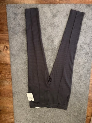 Mörkgrå randiga kostymbyxor - Snygga mörkgrå kostymbyxor med tunna vertikala ränder. Byxorna har en rak passform och klassisk midja. Materialet ser ut att vara en blandning av polyester och viskos för en dressad look. Perfekta för dig som vill ha en stilren och modern vibe.