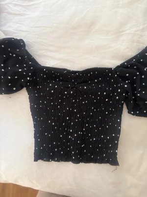 Svart prickig croppad topp - Trendig svart croppad topp med vita prickar och puffiga korta ärmar. Toppen har en smockad, stretchig passform och är perfekt för dig som gillar en söt och lekfull stil. Passar till både jeans och kjol.