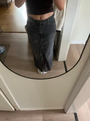 Mörkgrå lång jeanskjol med slits - Säljer en mörkgrå lång jeanskjol med hög midja och en snygg slits framtill. Kjolen har klassiska jeansdetaljer som fickor och knapp i midjan. Perfekt för dig som gillar en trendig och avslappnad stil.