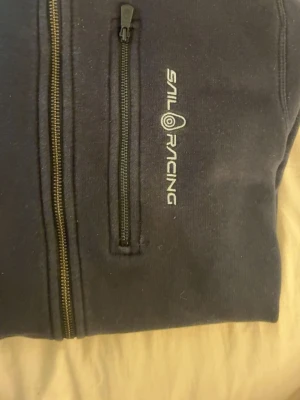 Marinblå hoodie från Sail Racing - Snygg marinblå hoodie från Sail Racing med dragkedja framtill och en bröstficka med zip. Hoodien har en klassisk huva och är tillverkad i mjukt material som känns skönt mot huden. Perfekt för en avslappnad och sportig stil.