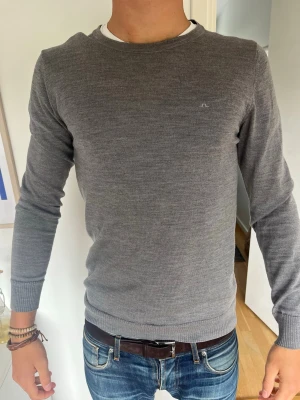 Grå tunnstickad tröja - Snygg grå tunnstickad tröja med rund halsringning och diskret logga på bröstet. Tröjan har långa ärmar och ribbade muddar vid ärmslut och nederkant. Model 185 cm, 71 kg bär S. Hör av er vid frågor