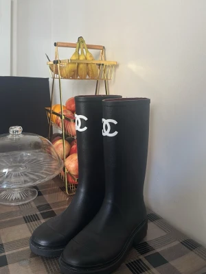 Chanel boots - Snygga svarta höga gummistövlar med vit logga framtill. Stövlarna har en klassisk, stilren design med rund tå och grov sula. Tillverkade i gummi som står emot regn och slask. Perfekta för en trendig look under regniga dagar.