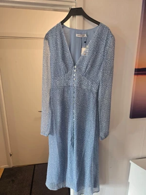 Blå blommig långklänning med knappar - Söt blå långklänning med små vita blommor, v-ringning och knappar framtill. Klänningen har lång ärm och är tillverkad i ett lätt, luftigt material som passar perfekt för vår och sommar.
