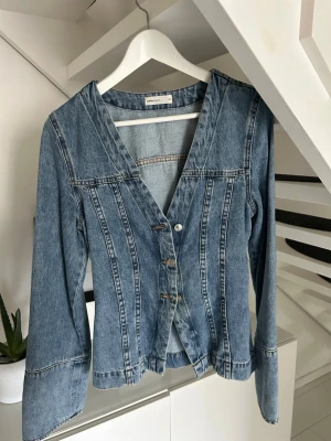 jeansjacka med v-ringning - Snygg jeansjacka i klassisk blå denim med markerade sömmar och v-ringad hals. Jackan har långa ärmar, knäppning framtill med fyra knappar och figurnära passform. Perfekt för dig som gillar en retro och unik stil.