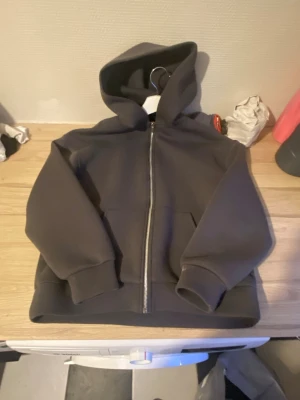 Mörkgrå hoodie med dragkedja - Säljer en stilren mörkgrå hoodie med dragkedja framtill och två fickor. Hoodien har en klassisk huva och breda muddar vid ärmar och nederkant. Perfekt för dig som gillar en enkel och clean look. Materialet känns mjukt och bekvämt.