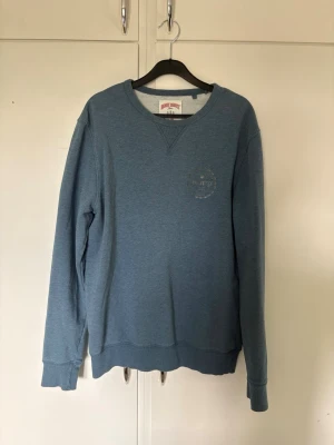 Tröja från Henry Choice  - En stilren blå crewneck sweatshirt med rund halsringning och ribbade muddar vid ärmar och nederkant. Tröjan har en diskret logga på bröstet och är tillverkad i mjukt material, perfekt för en avslappnad look. Bra skick 
