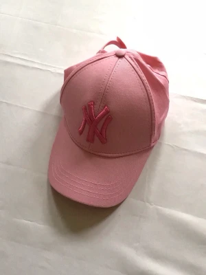 Rosa New York Yankees keps - Snygg rosa keps från New York Yankees med broderad NY-logga framtill i matchande rosa nyans. Klassisk böjd skärm och justerbar passform bak. Perfekt accessoar för en sportig och trendig look.