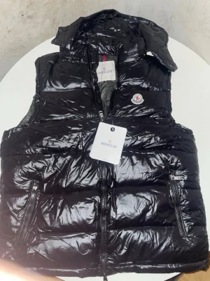 Svart glansig dunväst från Moncler - Snygg svart dunväst från Moncler med glansig finish och klassisk Moncler-logga på bröstet. Västen har dragkedja framtill, två fickor med dragkedja och hög krage. Perfekt för lager-på-lager och streetstyle.