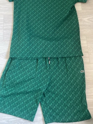 Grönt set från Lacoste med mönster - Säljer ett grönt set från Lacoste med matchande t-shirt och shorts. Setet har ett diskret geometriskt mönster över hela tyget och klassisk Lacoste-logga på shortsen. Perfekt för en avslappnad och sportig stil.