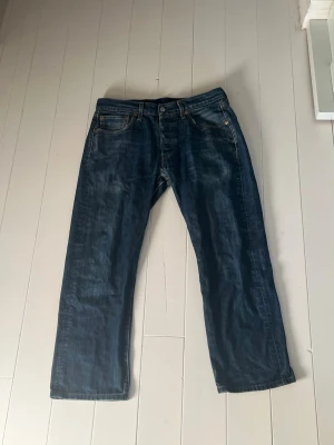 Raka Levi’s 501 jeans - Säljer ett par klassiska mörkblå jeans med rak/lös passform och fem fickor. Storlek W30 L30. Jeansen har kopparfärgade nitar och kontrastsömmar. Har dock skador mellan benen, syns på bild 5. Tillverkade i slitstarkt denimtyg, perfekta för en avslappnad stil. Väldigt snygg tvätt på den också som ger vintage känslan. Jeansen kan ha krympt lite.