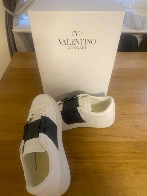 Valentino Garavani vita sneakers - Snygga vita sneakers från Valentino Garavani med bred svart rem över vristen och diskreta nitar. Skorna har en klassisk låg siluett och är tillverkade i läder för en exklusiv känsla. Perfekta för dig som vill ha en stilren och trendig look.