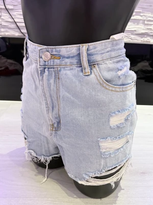 Ljusblå high waist jeansshorts - Säljer ett par ljusblå denimshorts med hög midja och slitna detaljer. Shortsen har fransiga benslut, flera revor framtill och klassisk femficksdesign. Perfekta för sommaren och ger en avslappnad vibe.
