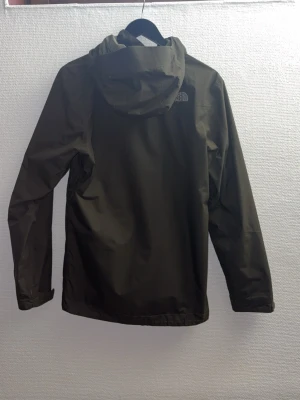 Olivgrön skaljacka från The North Face - Snygg olivgrön regnjacka från The North Face med huva, dragkedja och flera praktiska fickor. Jackan har en minimalistisk design och är tillverkad i ett vattenavvisande material, perfekt för regniga dagar. Logotyp på bröstet och ärmen.