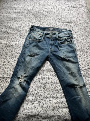 Blå slitna jeans från Replay - Säljer ett par blå jeans från Replay, modell Anbass. Jeansen har en slim fit passform, slitningar och hål på benen för en trendig, avslappnad look. Klassisk femficksmodell med dragkedja och knapp. Perfekta för dig som gillar en edgy stil.