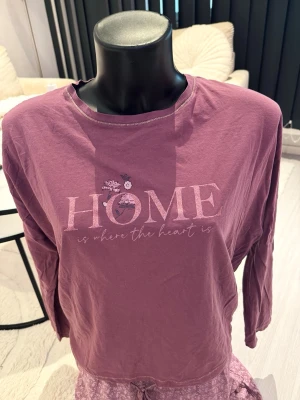 Lila pyjamas med tryck och mönstrade byxor - Mysig lila pyjamas med långärmad tröja med texten 'HOME is where the heart is' och små blommor. Byxorna har ett diskret mönster och dragsko i midjan. Perfekt för chillkvällar hemma.