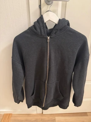 Mörkgrå zip hoodie med huva - En enkel och stilren mörkgrå hoodie med dragkedja framtill och två stora fickor. Hoodien har en klassisk huva och ribbade muddar vid ärmar och nederkant. Perfekt för chill dagar eller när du vill ha något bekvämt. Materialet känns mjukt och skönt.