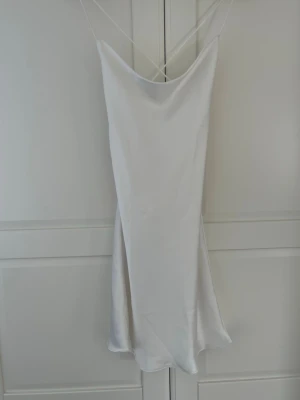 Vit satinklänning - Säljer en elegant vit klänning i satin med smala axelband som korsas i ryggen. Klänningen har en lös passform och en snygg draperad rygg. Perfekt för dig som gillar minimalistisk och stilren design. Aldrig använd!
