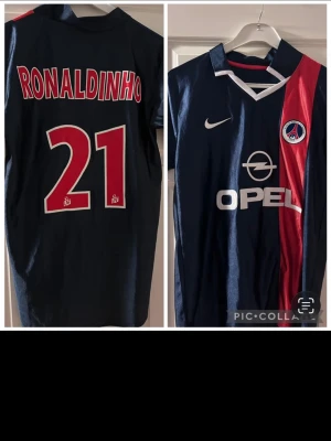 PSG fotbollströja Ronaldinho #21 - Säljer en klassisk PSG matchtröja från Nike med Ronaldinho och nummer 21 på ryggen. Tröjan är mörkblå med röd och vit detalj, har klubbmärke och Opel-logga på bröstet samt V-ringad krage. Perfekt för dig som älskar fotboll och retrostil.