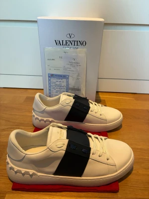Valentino Garavani open sneakers - Snygga vita sneakers från Valentino Garavani med bred svart rem över vristen och coola nitar runt hälen. Skorna är i läder och har vita snören samt diskret logga på plösen. Perfekta för dig som vill ha en stilren men edgy look.