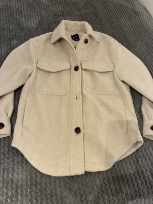 Beige overshirt jacka från Bershka storlek xxs - Stilren beige overshirt jacka från Bershka i mjukt fleece-liknande material. Två stora bröstfickor med lock, klassisk krage och knäppning med mörka knappar. Perfekt lager-på-lager-plagg för en avslappnad look. Fickor i sidorna