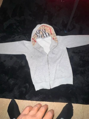 Grå zip hoodie med mönstrad huva - Säljer en grå zip hoodie från Burberry med klassiskt rutmönster i huvan och broderad logga på bröstet. Hoodien har dragsko i huvan, fickor framtill och ribbade muddar. Perfekt för en chill och stilren look.