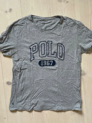 Grå POLO t-shirt från Ralph Lauren - Snygg grå t-shirt från Polo Ralph Lauren med stor mörkblå POLO-text och siffran 1967 på bröstet. Tillverkad i mjuk bomull och har en klassisk rund hals. Perfekt för en avslappnad och stilren look.
