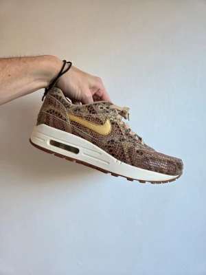 Nike Air Max 1 ormmönstrade sneakers - Unika Nike Air Max 1 sneakers med ormmönstrad ovandel i bruna och gröna toner. Guldigt swoosh på sidan och vit mellansula med synlig Air-enhet. Yttersulan är brun och skorna har klassisk snörning. Materialet är en mix av textil och syntet.