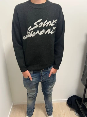 Svart saint Laurent hoodie - Lite stor på mig ja är 177 Snygg svart stickad tröja med vit text i handskriven stil över bröstet. Tröjan har rund halsringning och långa ärmar med ribbade muddar vid ärmslut och nederkant. Perfekt för dig som gillar streetwear och vill ha en trendig look.