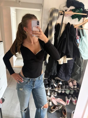 Svarr body med twistad detalj - Säljer en svart långärmad body med djup v-ringning och twistad detalj framtill. Bodyn har en åtsittande passform och är tillverkad i ett stretchigt material. Köpt second hand och uppskattar storlek S. Har inget märke.
