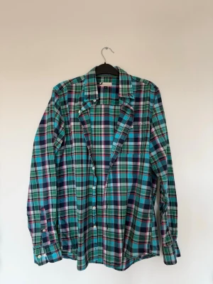 Grön/blå rutig skjorta i storlek M - Rutig långärmad skjorta med knappar framtill och på ärmsluten. Skjortan har en bröstficka på vänster sida. Ger lite mer färg till outfiten och fungerar väldigt bra både oversize och öppet.