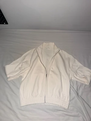 Vit zip-jacka från Zara  med resårdetaljer - Stilren vit jacka från Zara med dragkedja framtill och resår vid ärmslut och midja. Jackan har hög krage och raka ärmar, perfekt för en clean och enkel look. Passar till det mesta och är lätt att matcha med andra plagg. Har använts en gång. Priset kan diskuteras. 
