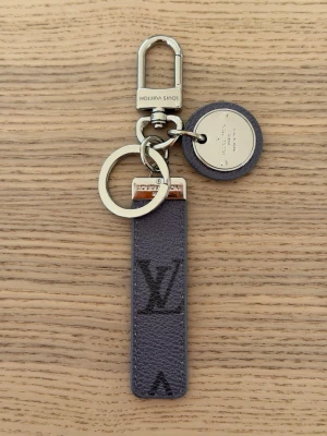 Vuitton nyckelring i grått läder - Snygg nyckelring från  Vuitton i grått läder med metalliska detaljer och LV-logga. Har både rund bricka och klassisk karbinhake i silverfärg. Perfekt accessoar för att piffa upp nyckelknippan eller väskan.