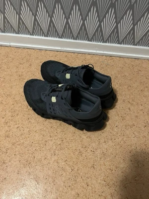 Svarta sneakers med mesh och snabbsnörning - Säljer ett par svarta ON sneakers med sportig design och snabbsnörning. Ovandelen är i mesh och syntetmaterial för bra andningsförmåga. Sulan är kraftig och ger bra grepp. Perfekta för dig som gillar en aktiv stil.