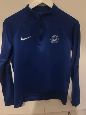 Blå PSG träningströja från Nike - Snygg blå långärmad träningströja från Nike med PSG-logga på bröstet. Tröjan har halv dragkedja framtill och är tillverkad i ett stretchigt funktionsmaterial som passar perfekt till träning eller uppvärmning.