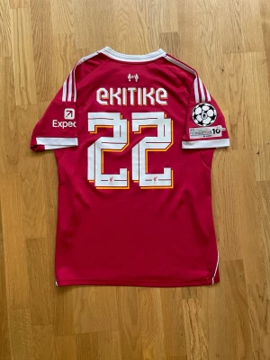 Ekitike #22 - S - Säljer en röd Liverpool matchtröja från Adidas med vita detaljer och tryck. Tröjan har nummer 22 och namnet Ekitike på ryggen, Champions League-märke på ärmen och sponsorloggor på bröstet. Perfekt för dig som älskar fotboll och vill sticka ut på planen.