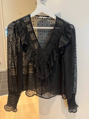 Svart volangblus från ONLY - Säljer en svart transparent blus från ONLY i storlek XS. Blusen har volangdetaljer framtill, v-ringning, knappar och långärmade ärmar med smock vid ärmslut. Tunt tyg med diskret prickigt mönster för en trendig look.