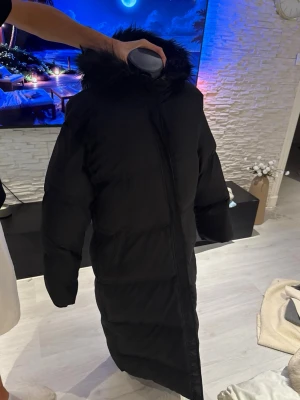 Lång svart dunjacka med huva - Säljer en lång svart dunjacka med stor huva och fejkpäls runt kanten. Jackan har en rak passform och dragkedja framtill. Perfekt för kalla vinterdagar när du vill hålla dig varm och stilsäker. Klassisk design som funkar till det mesta.