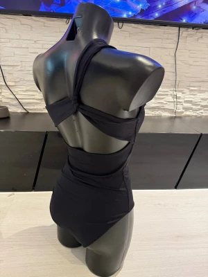 Svart oneshoulder baddräkt - Snygg svart baddräkt med oneshoulder-design och öppen rygg. Baddräkten har en modern och stilren look med bred axelrem och coola utskärningar i ryggen. Perfekt för dig som vill sticka ut på stranden eller vid poolen.