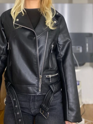 Svart skinnjacka - Snygg svart skinnjacka med silvriga dragkedjor, breda slag och bälte i midjan. Jackan har flera fickor med dragkedja och en cool bikerstil som passar perfekt till många outfits. Helt ny med prislapp kvar. (Nypris 499kr) 