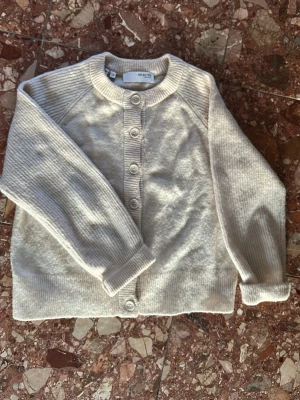 Beige kofta från Selected Femme - Mysig beige kofta från Selected Femme med rund halsringning och knäppning framtill. Koftan har långa ärmar, ribbade muddar och är stickad i ett mjukt material. Perfekt för lager-på-lager och enkel att matcha med olika outfits.