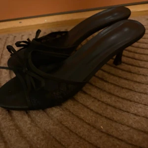 Svarta spetsiga pumps med rosett - Säljer ett par svarta pumps med öppen tå och liten klack. Skorna har en elegant spetsdetalj och en söt rosett framtill. Materialet ser ut att vara en mix av textil och syntet, vilket ger en stilren look. Perfekta för dig som vill ha något klassiskt och snyggt. Storlek 39. Använd få gånger.