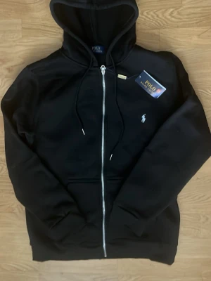 Svart hoodie från Polo Ralph Lauren - Snygg svart hoodie med dragkedja från Polo Ralph Lauren. Klassisk design med huva och justerbara snören, samt den ikoniska vita logon broderad på bröstet. Tillverkad i mjukt material som passar perfekt till en avslappnad stil.