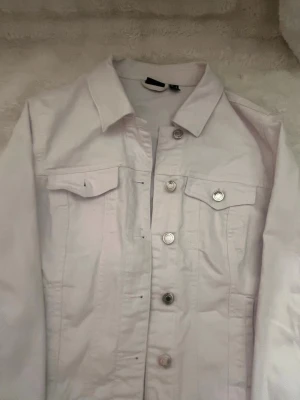 Vit jeansjacka från Vero Moda - Snygg vit jeansjacka från Vero Moda med klassisk krage, silverfärgade knappar och två bröstfickor med lock. Jackan har en rak passform och är perfekt för att lyfta vilken outfit som helst. Enkel och stilren design som passar till mycket.